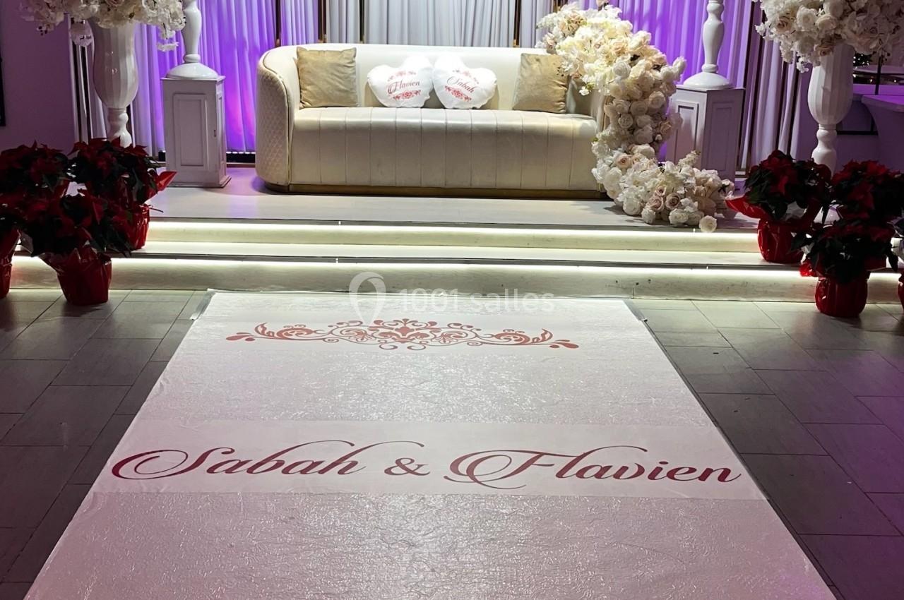 Décor de mariage avec canapé beige, fleurs blanches et tapis personnalisé portant des prénoms en rouge.