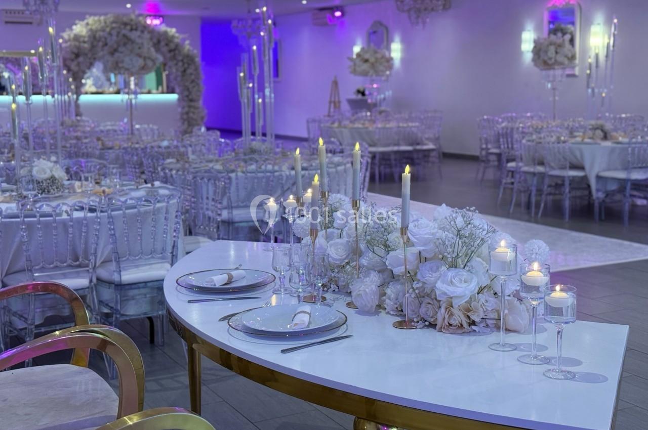 Salle de réception décorée avec des tables élégantes, des chandeliers et des arrangements floraux blancs.