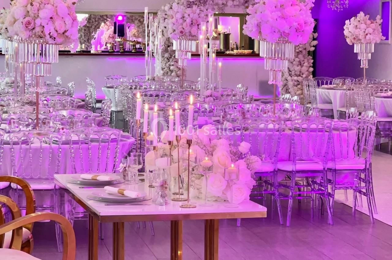 Salle de réception décorée avec des tons roses, chandeliers, fleurs blanches et tables élégamment dressées.