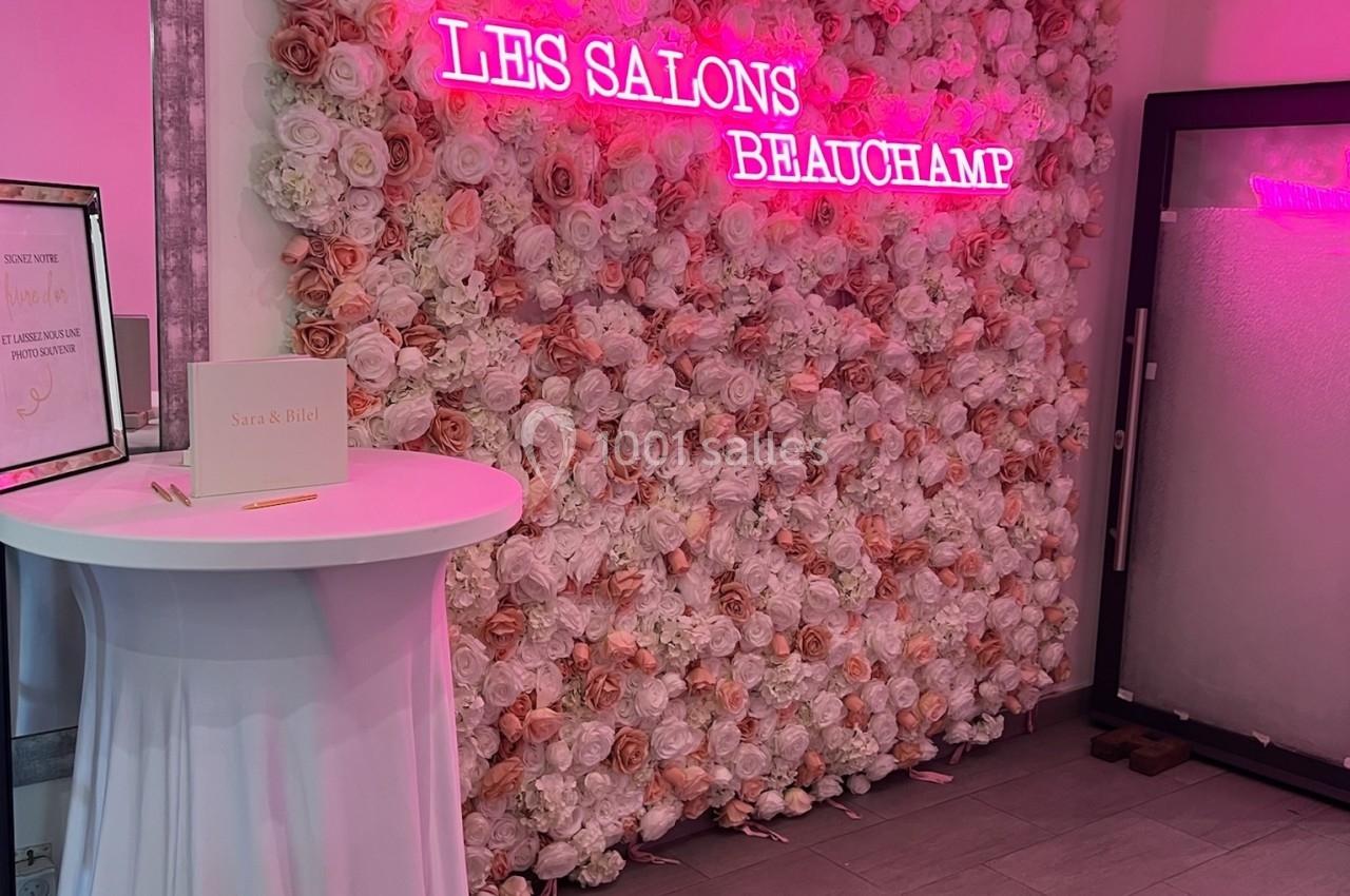 Mur de fleurs roses et blanches avec une enseigne néon rose ’Les Salons Beauchamp’, table haute drapée à gauche.