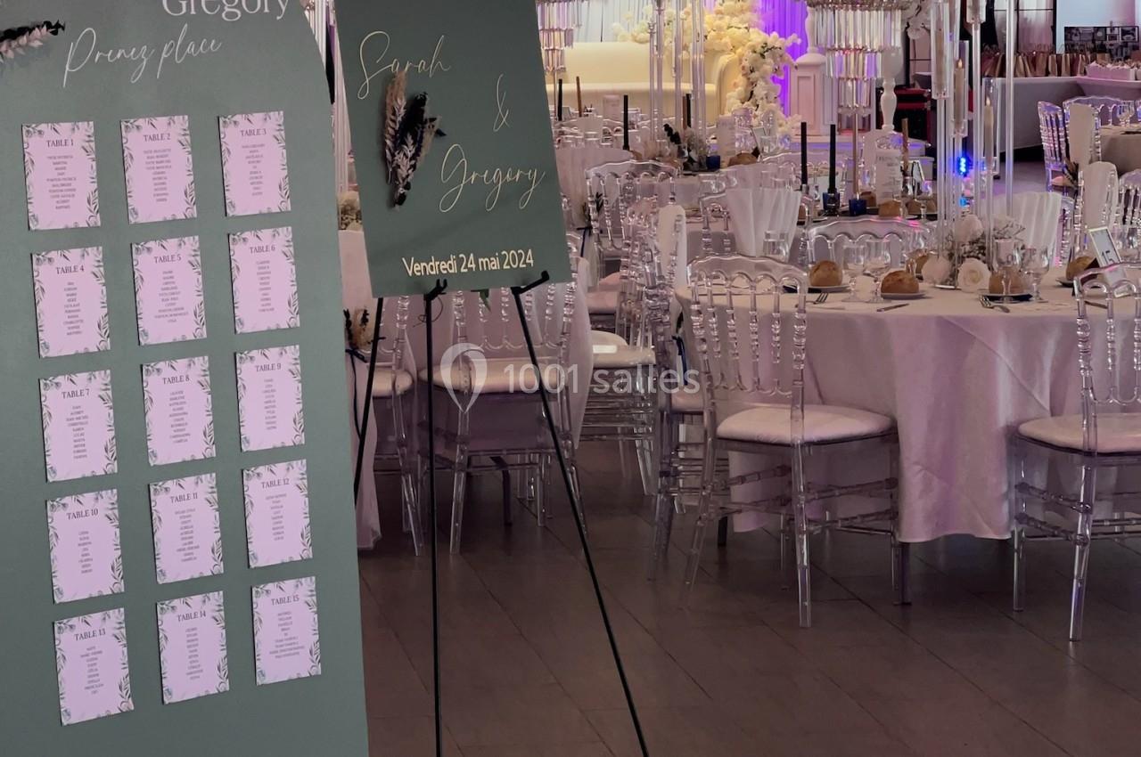Salle de réception décorée pour un mariage avec panneaux de bienvenue et plan de table, tables dressées et fleurs blanches.