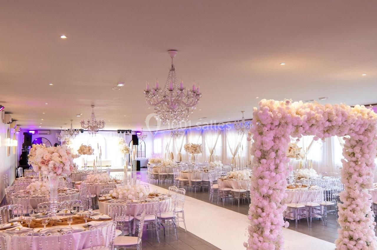 Salle de réception décorée pour un mariage avec tables dressées, chandeliers et arche florale blanche.