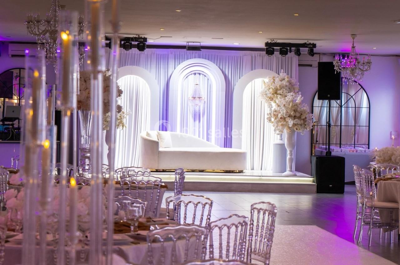 Salle de réception élégante avec décoration florale blanche, chandeliers et canapé devant des arches lumineuses.