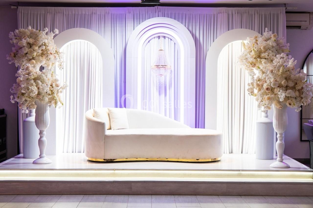 Canapé blanc sur une scène décorée de rideaux, arches lumineuses et arrangements floraux blancs.