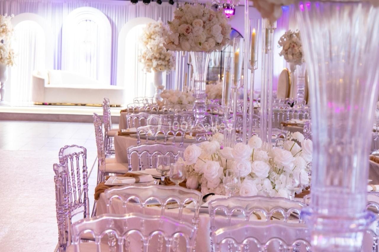 Salle de réception élégante avec tables décorées de fleurs blanches, chaises transparentes et éclairage tamisé.