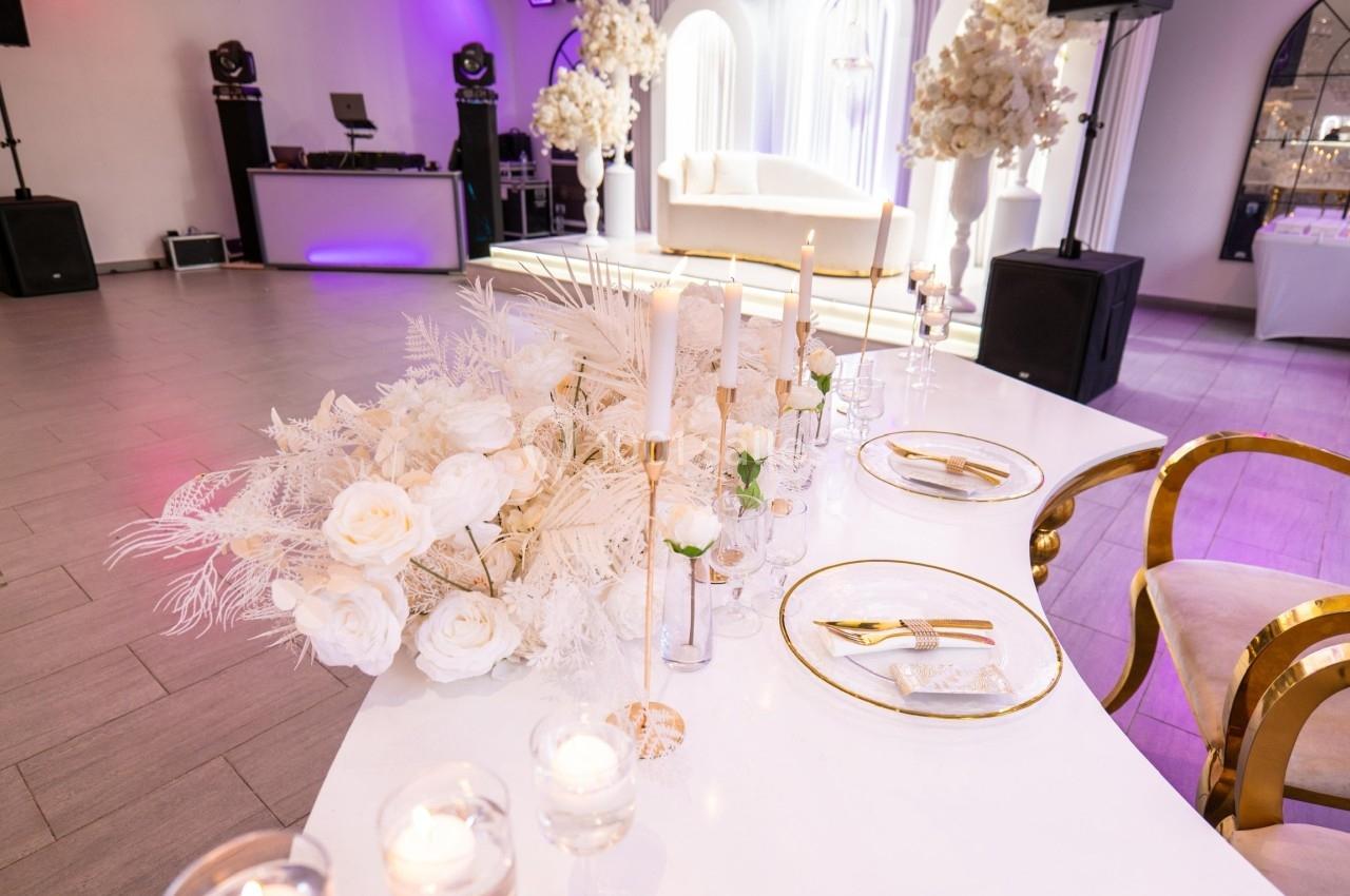 Table élégamment décorée avec des fleurs blanches, bougies et vaisselle dorée dans une salle de réception lumineuse.