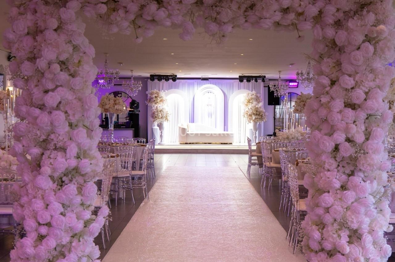 Arche florale blanche encadrant une allée menant à une scène décorée pour un événement festif en intérieur.