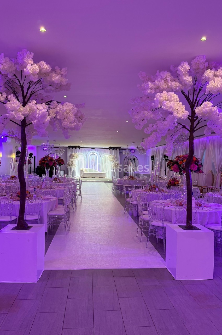 Salle de réception décorée avec des arbres fleuris, des tables dressées et une allée centrale menant à une estrade éclairée.