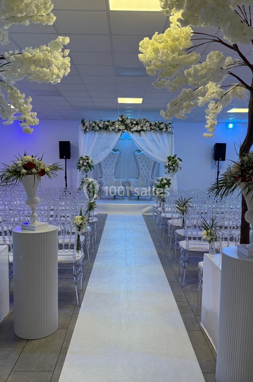 Allée centrale décorée de fleurs blanches menant à une arche ornée pour une cérémonie dans une salle lumineuse.