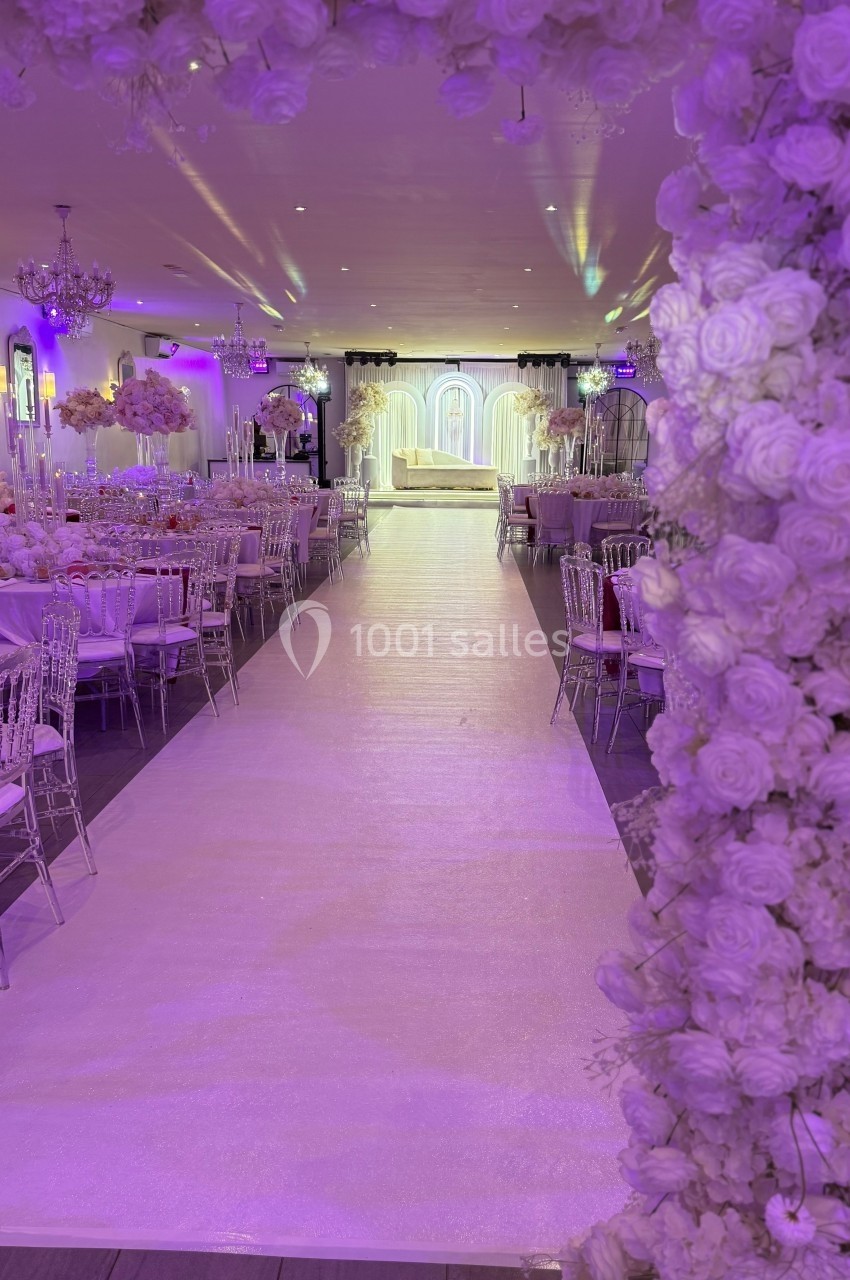 Allée centrale décorée de fleurs blanches menant à une scène illuminée dans une salle de réception élégante.