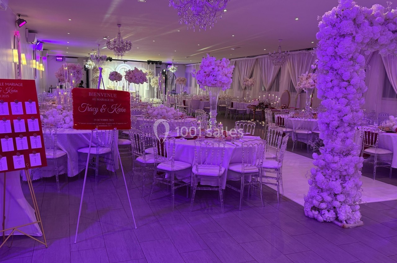 Salle de réception décorée pour un mariage avec tables dressées, arche florale blanche et plan de table affiché.