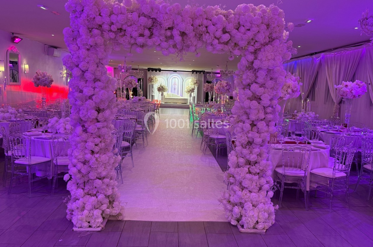 Allée centrale décorée de fleurs blanches menant à une arche dans une salle de réception éclairée de tons violets.