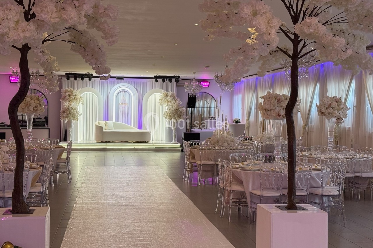 Salle de réception élégante décorée de fleurs blanches, avec une allée centrale menant à une scène éclairée.