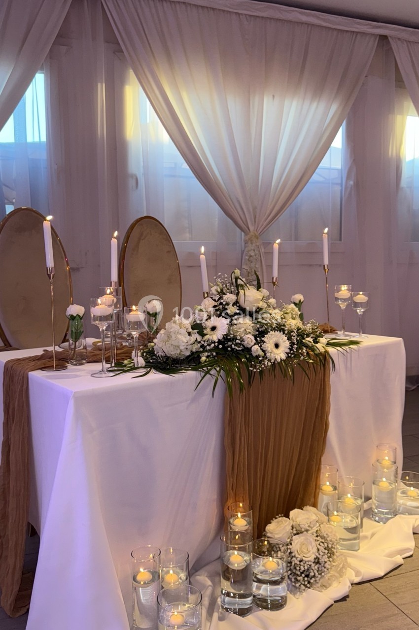 Table décorée pour un mariage avec fleurs blanches, bougies, nappes blanches et rideaux translucides en arrière-plan.