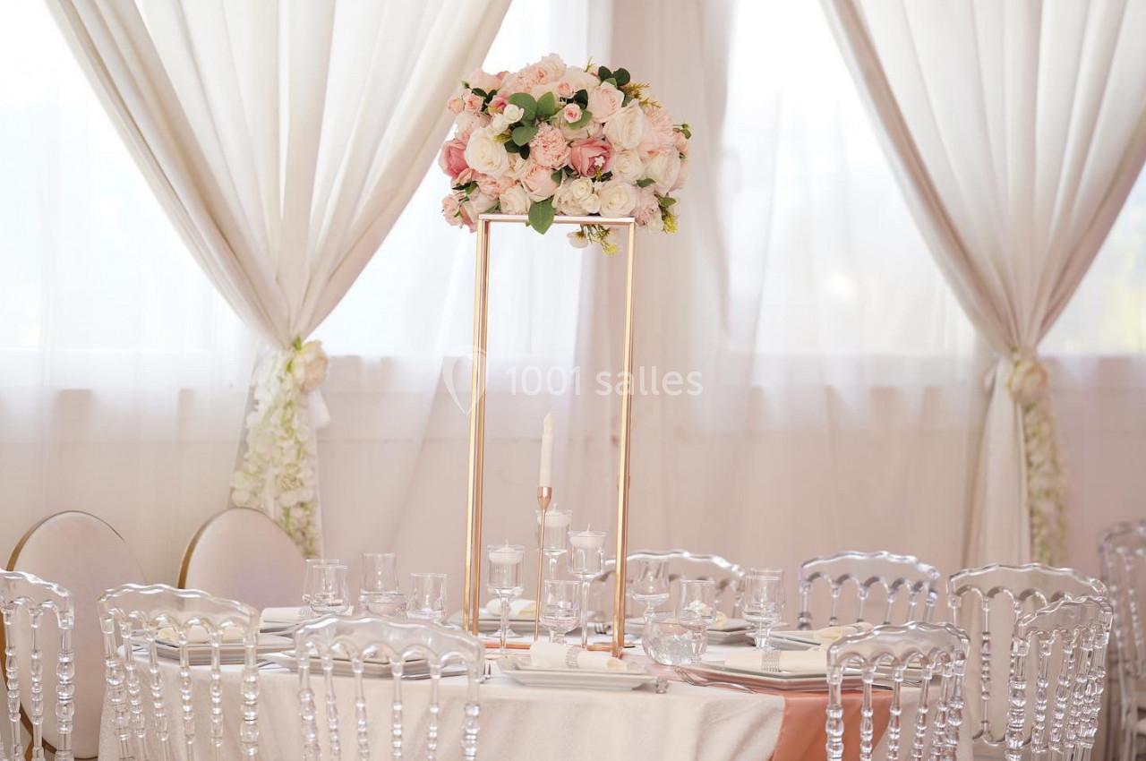 Centre de table floral avec roses blanches et roses sur un support doré, entouré de chaises transparentes et nappes blanches.