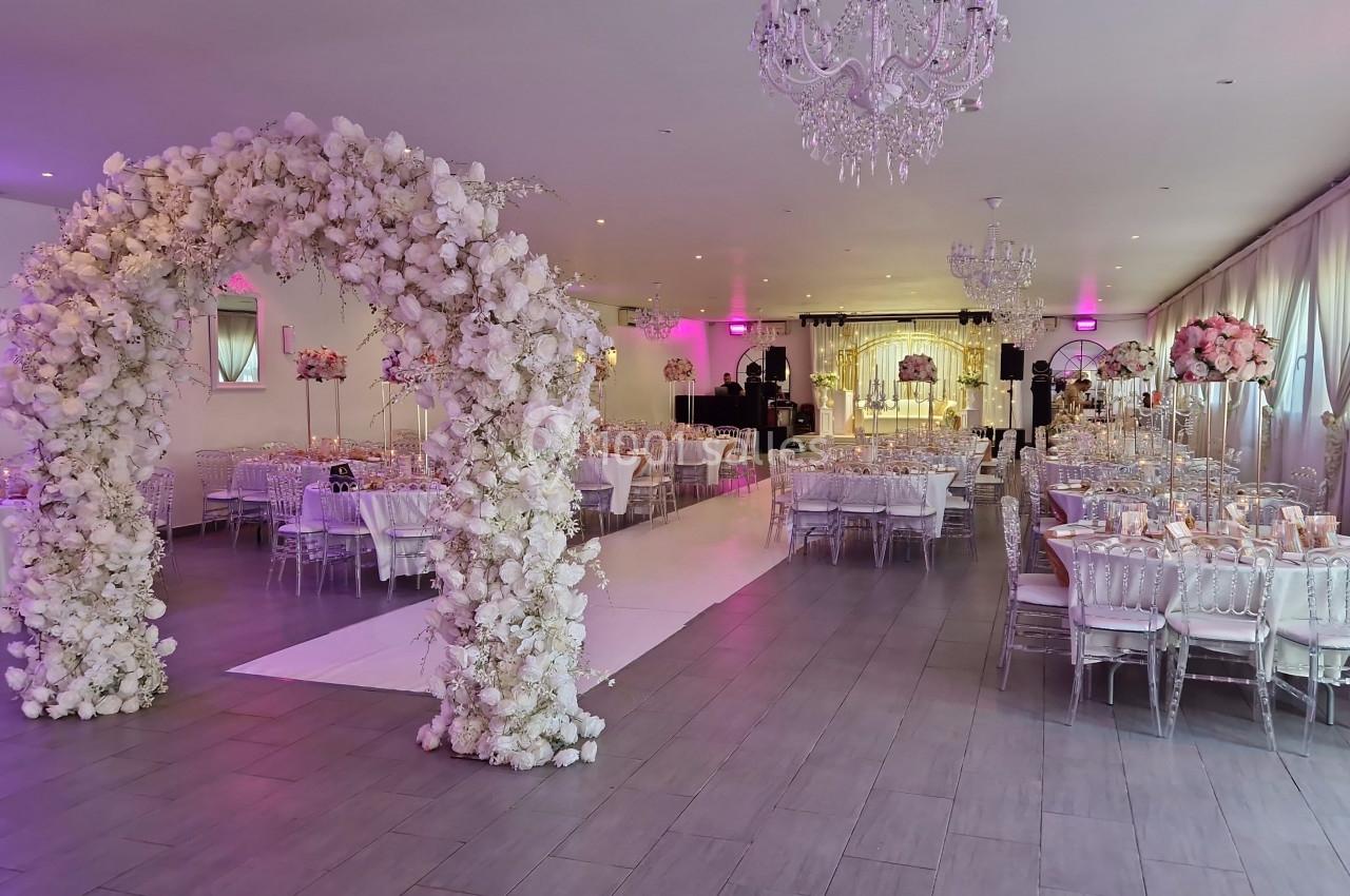 Salle de réception décorée avec des tables élégantes, un arche floral blanc et un éclairage rose tamisé.