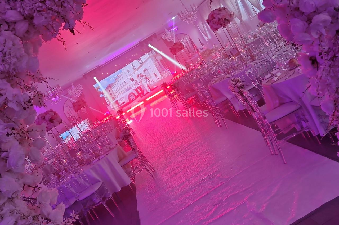Salle de réception décorée avec des fleurs blanches, des tables dressées et un éclairage rose dominant.