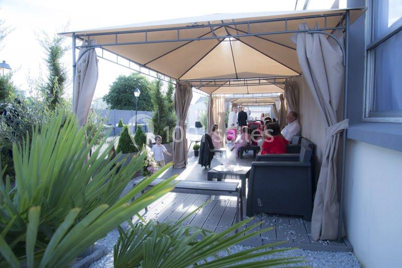 Des personnes assises et debout sous des pergolas avec rideaux, dans un espace extérieur aménagé avec des plantes.