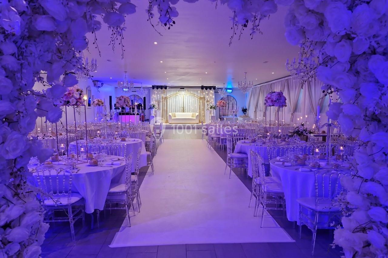 Salle de réception décorée avec des fleurs blanches, tables élégantes et éclairage bleu, centrée sur une estrade.
