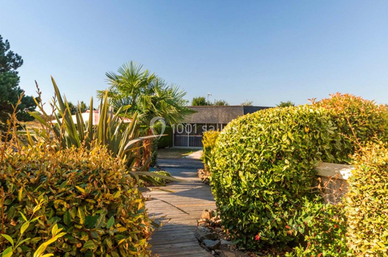 Allée en bois bordée de buissons et de plantes menant à une maison moderne sous un ciel dégagé.