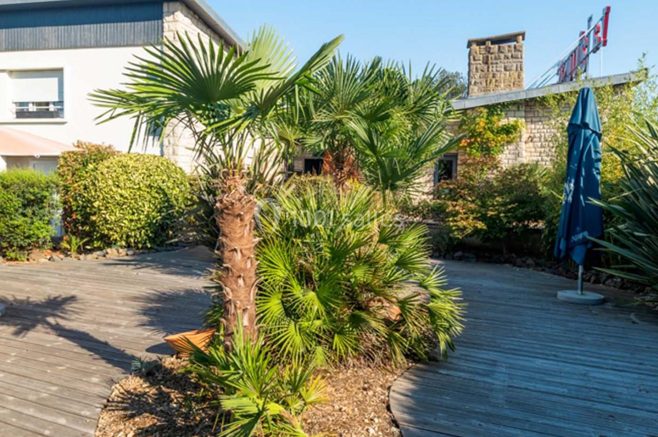 Terrasse en bois entourée de palmiers et de végétation, avec un parasol bleu et un bâtiment en arrière-plan.