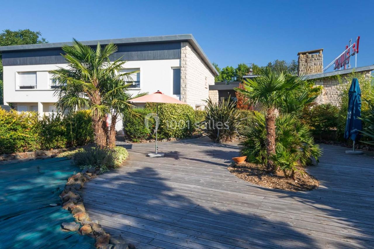 Jardin avec terrasse en bois, palmiers et parasols devant un bâtiment moderne sous un ciel bleu.