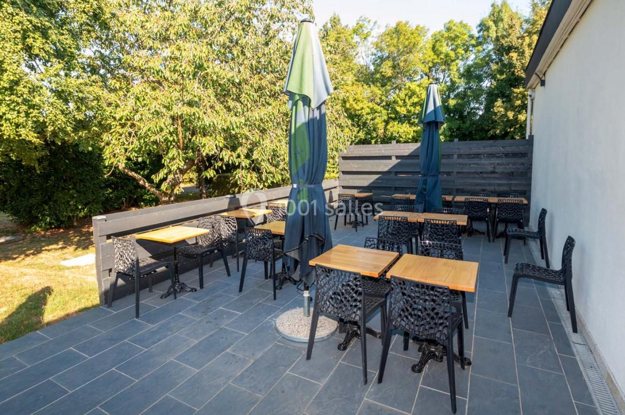 Terrasse extérieure avec tables en bois, chaises noires et parasols, entourée de verdure et d'une clôture en bois.