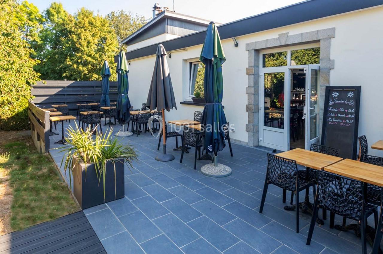 Terrasse extérieure d'un restaurant avec tables, chaises et parasols, entourée de verdure et d'un bâtiment clair.