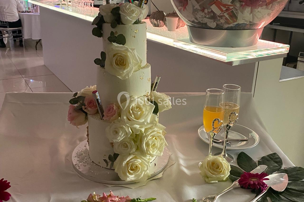 Gâteau de mariage à trois étages décoré de roses blanches et roses, posé sur une table avec des fleurs et des coupes.