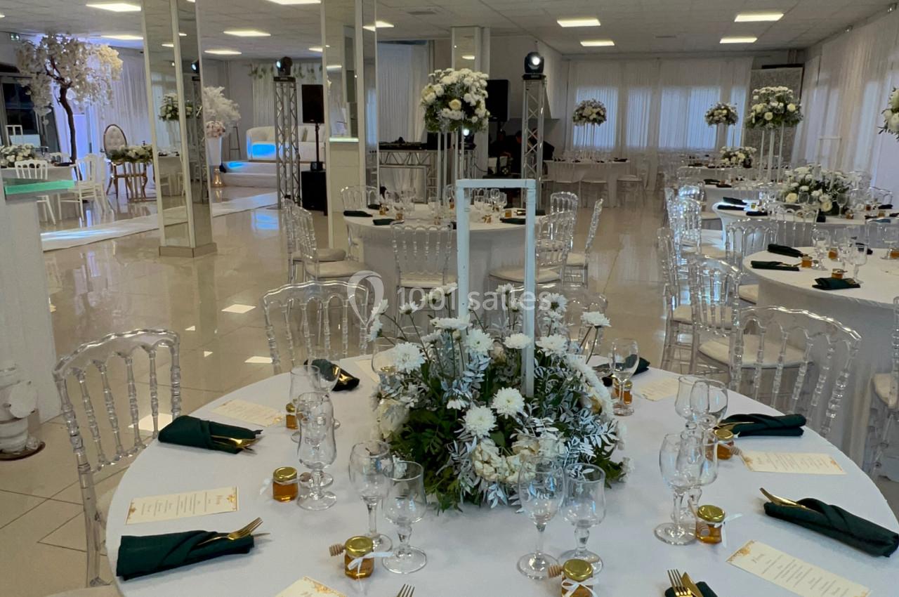 Salle de réception lumineuse avec tables rondes décorées de nappes blanches, centres de table floraux et serviettes vertes.