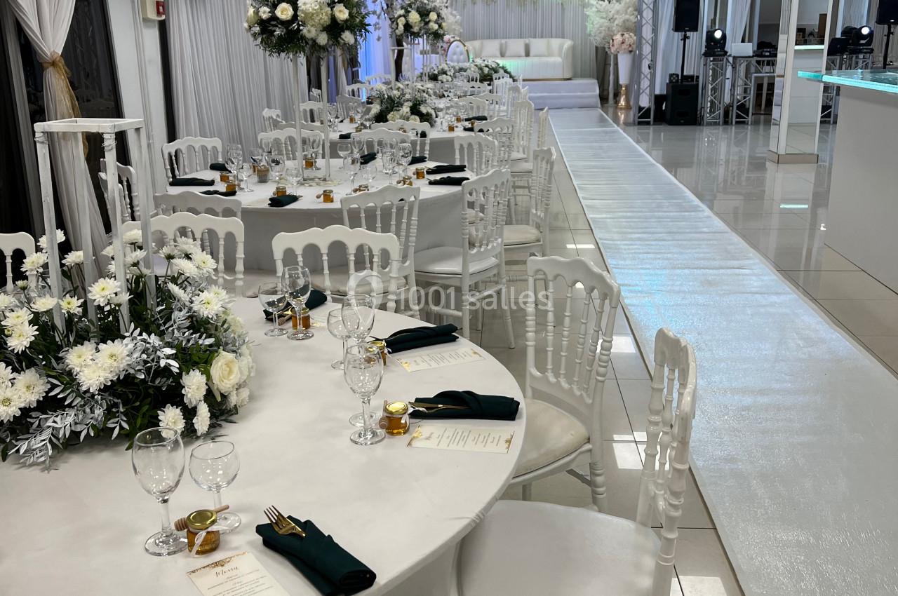 Salle de réception décorée avec des tables rondes, nappes blanches, chaises blanches et arrangements floraux élégants.