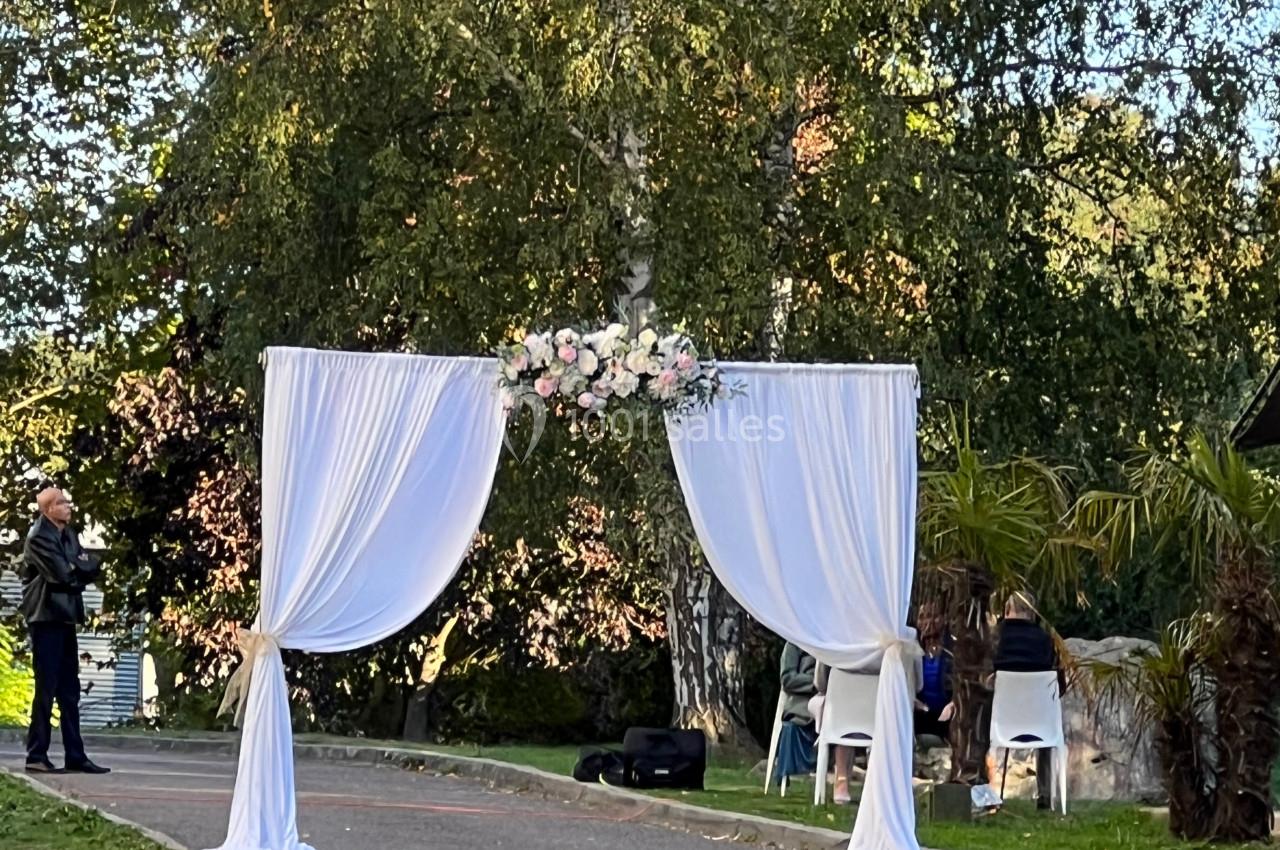 Arche de mariage décorée de fleurs blanches et roses, installée en extérieur sur une pelouse bordée d'arbres.