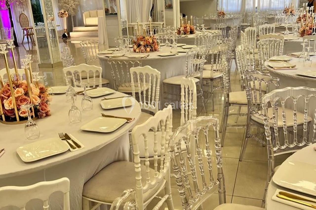 Salle de réception décorée avec des tables rondes dressées, chaises transparentes et arrangements floraux élégants.