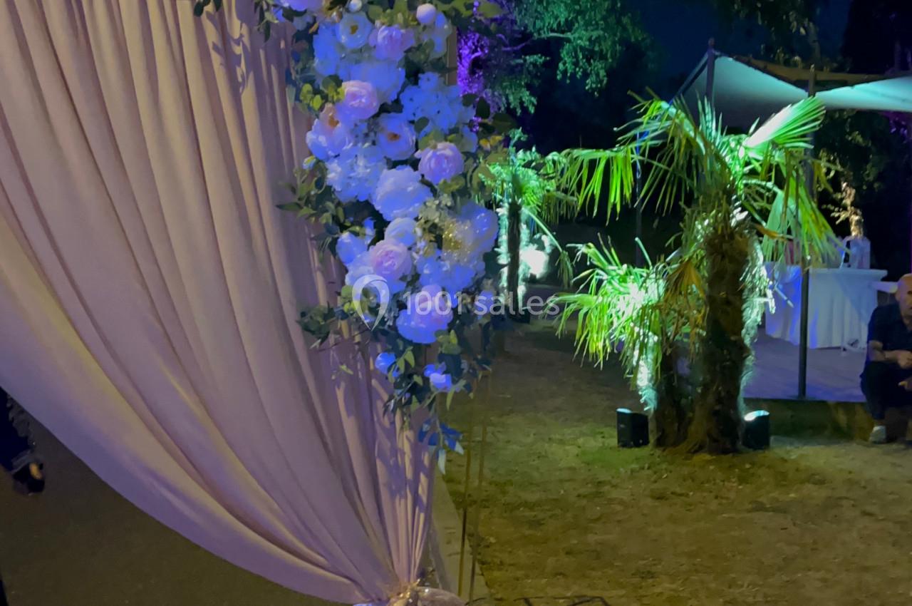 Rideau beige noué décoré de fleurs blanches, installé en extérieur de nuit avec des arbres illuminés en arrière-plan.