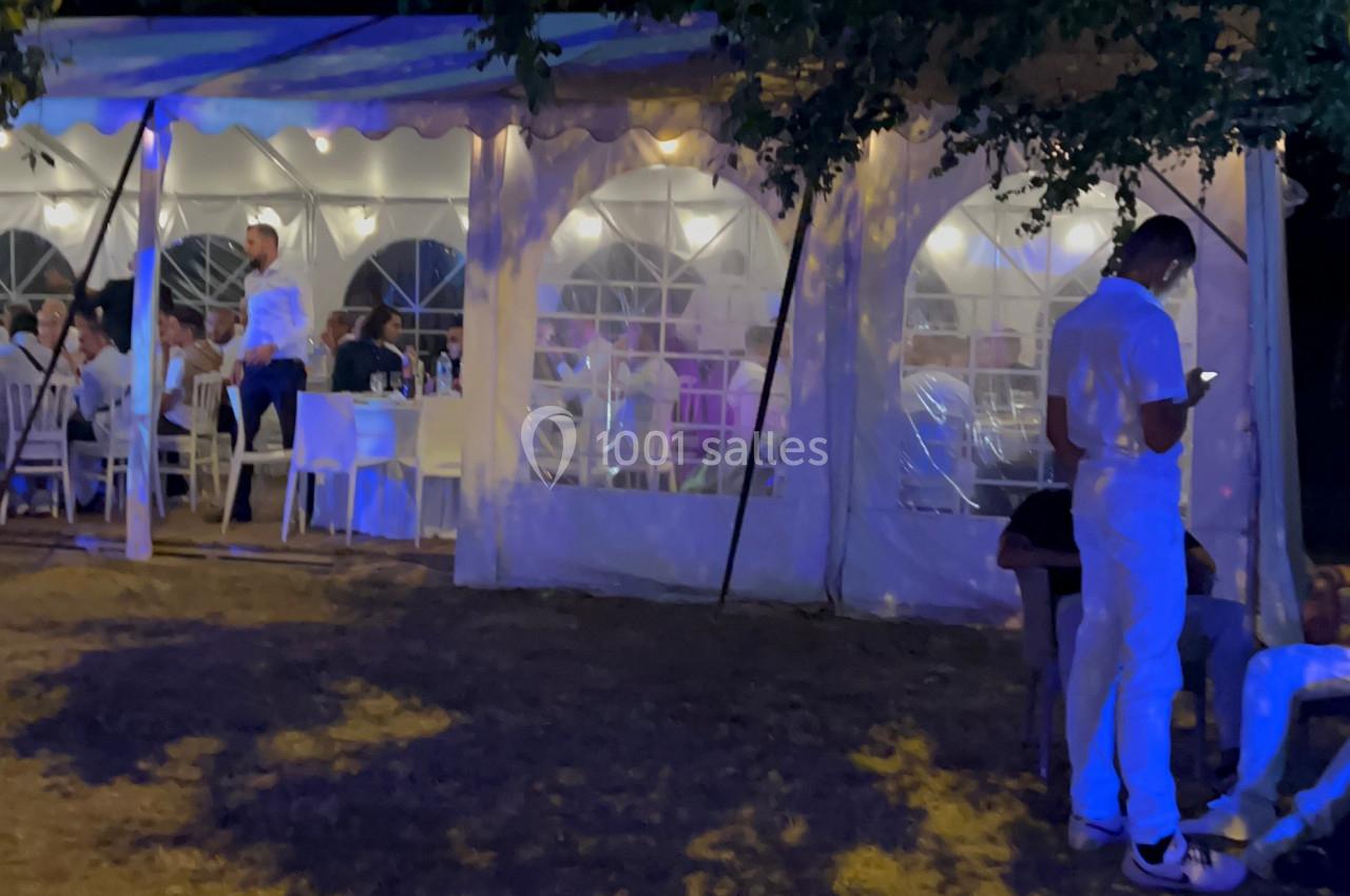 Un chapiteau éclairé en soirée avec des personnes assises et debout, entouré d'arbres et d'ombres au sol.