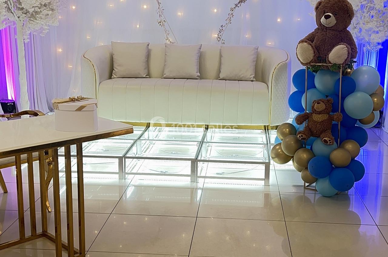 Canapé blanc devant un rideau lumineux en forme de cœur, décoré avec ballons, ours en peluche et arbres artificiels.