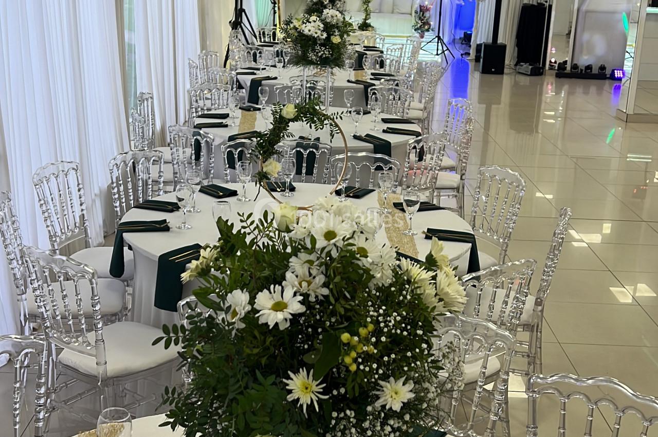 Salle de réception décorée avec des tables dressées, nappes blanches, chaises transparentes et centres de table fleuris.