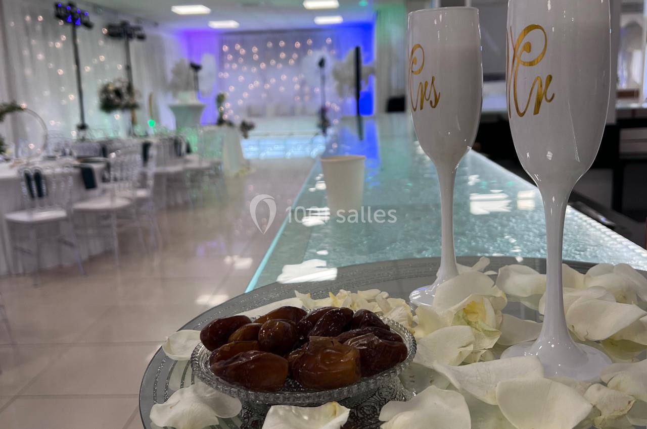 Plateau avec des dattes, pétales de rose et flûtes décorées sur un comptoir, salle de réception en arrière-plan.
