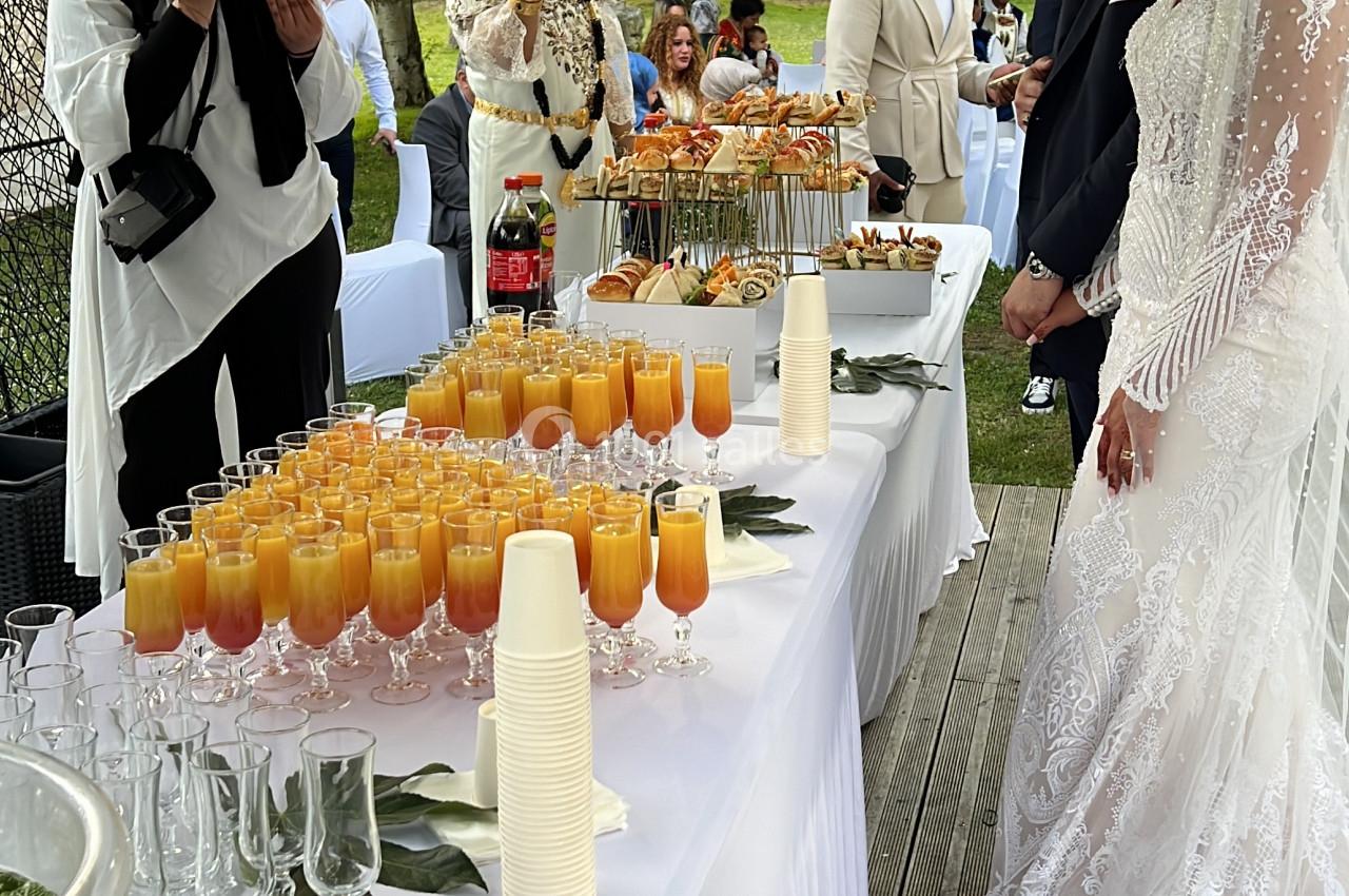 Un couple en tenue de mariage se tient près d'une table de buffet décorée, entouré d'invités et de photographes.