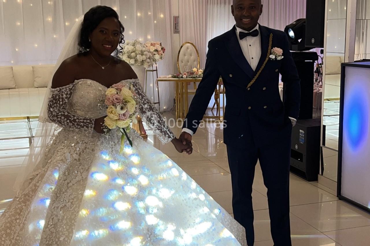 Un couple en tenue de mariage pose dans une salle lumineuse avec une robe illuminée et un décor élégant.