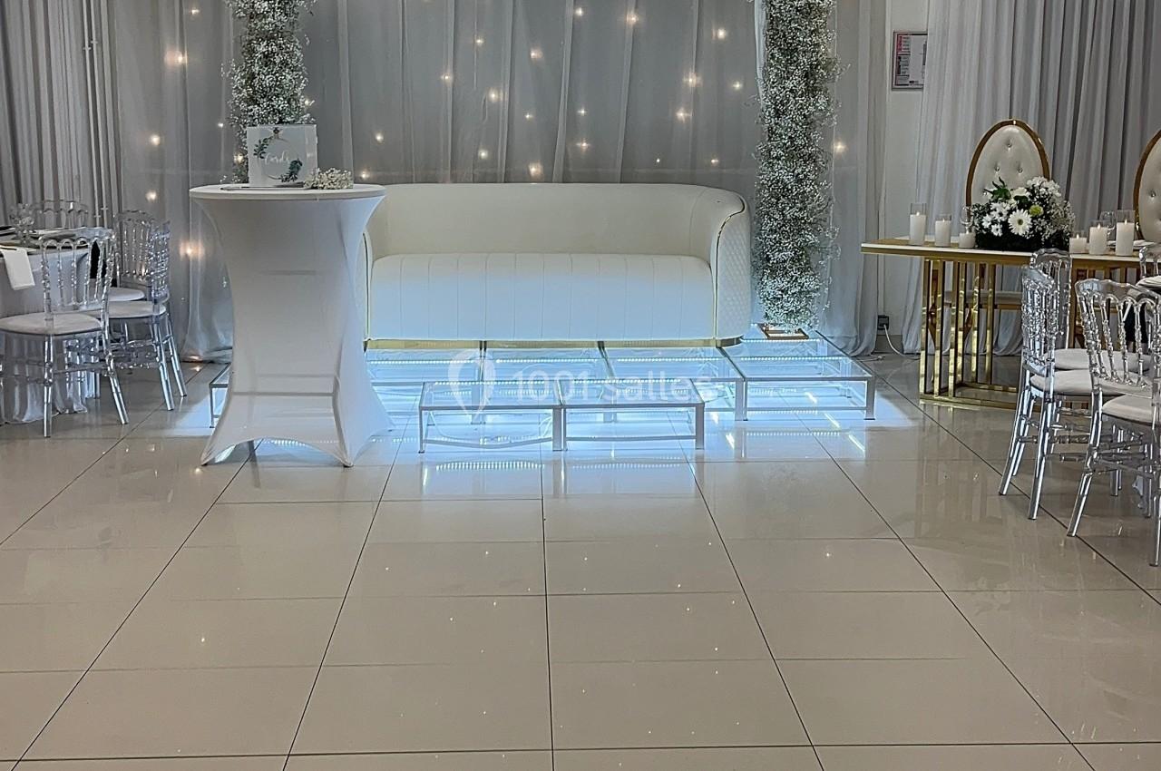 Salle de réception lumineuse avec canapé blanc, arches fleuries et tables décorées en arrière-plan.