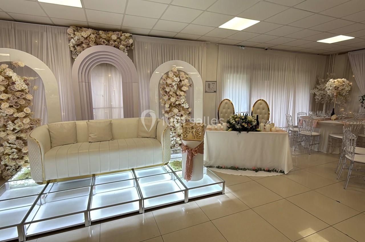 Salle de réception décorée avec des arches fleuries, un canapé blanc sur podium lumineux et des tables élégantes.