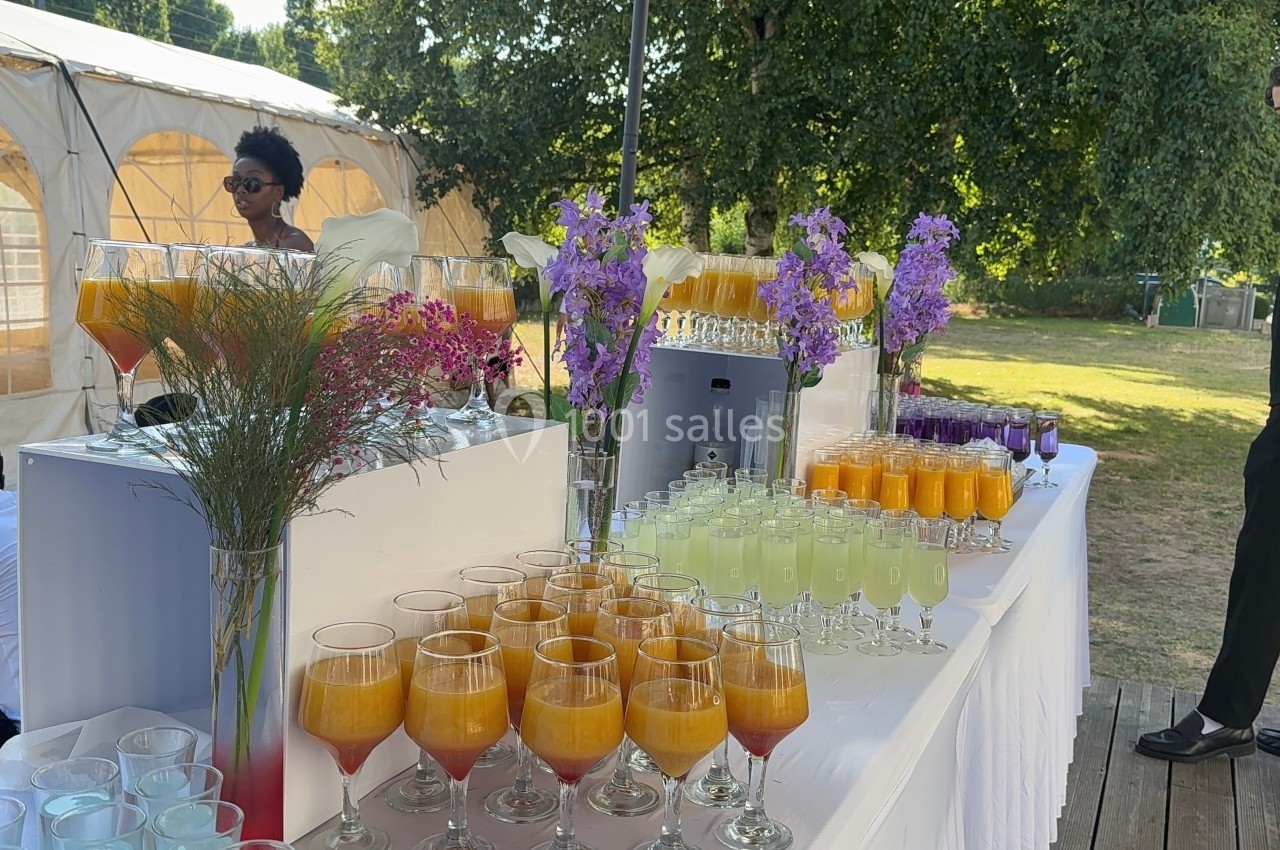 Table avec des verres de cocktails colorés, décorée de fleurs, installée en extérieur près d'une tente blanche.