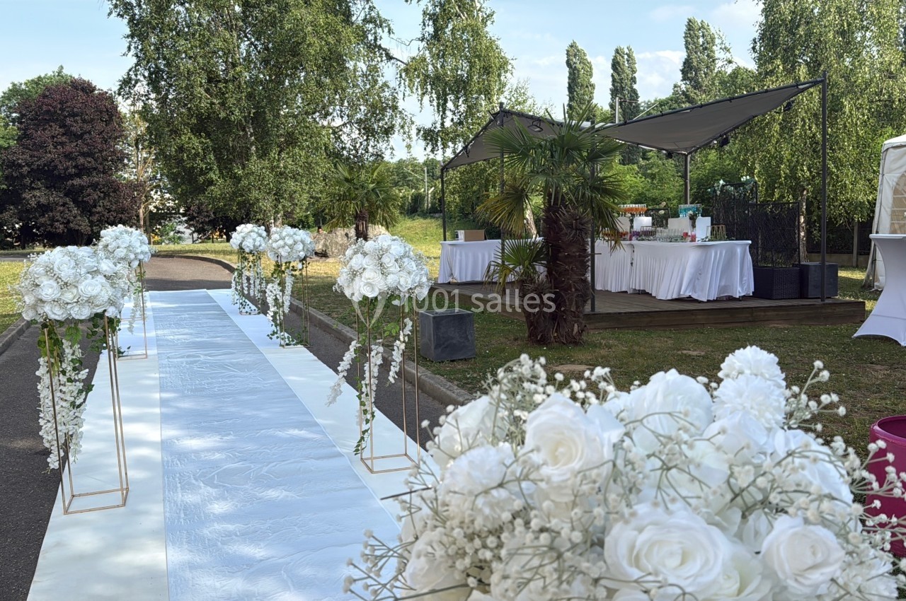 Allée décorée de fleurs blanches menant à un espace extérieur aménagé avec tables sous des tonnelles.