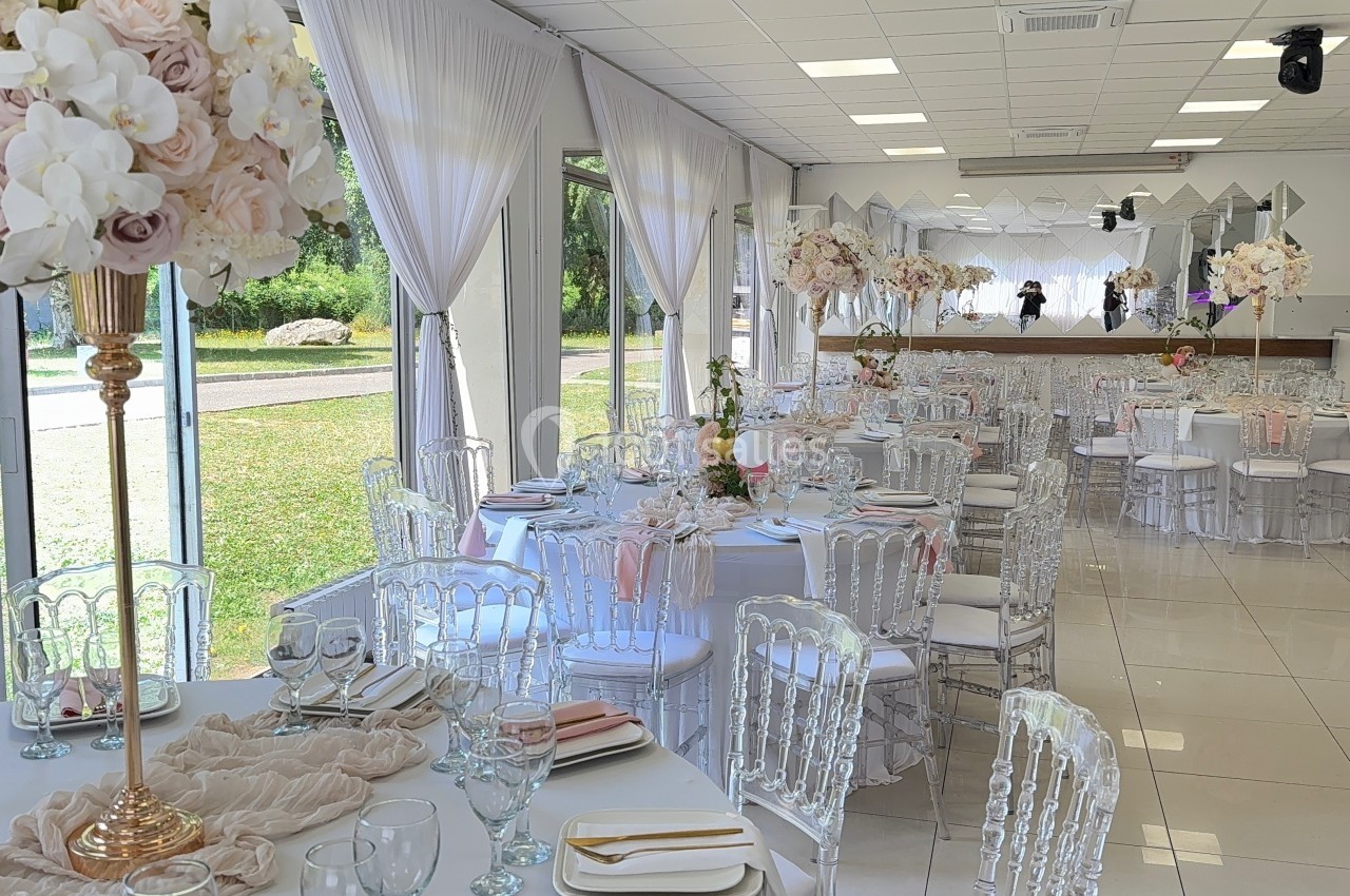 Salle de réception lumineuse avec tables décorées de nappes blanches, chaises transparentes et centres de table fleuris.