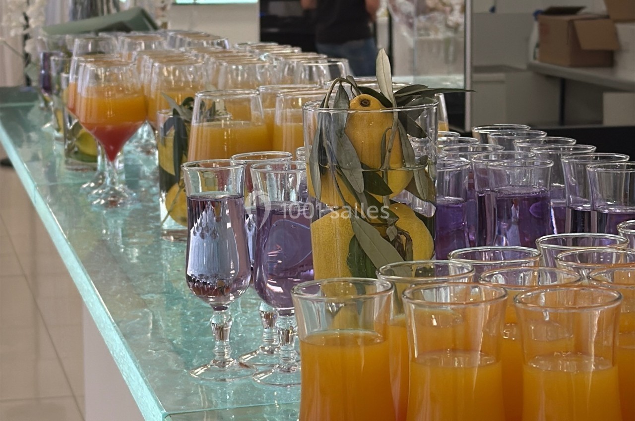 Verres de cocktails colorés alignés sur un comptoir en verre, avec des fleurs blanches en arrière-plan.