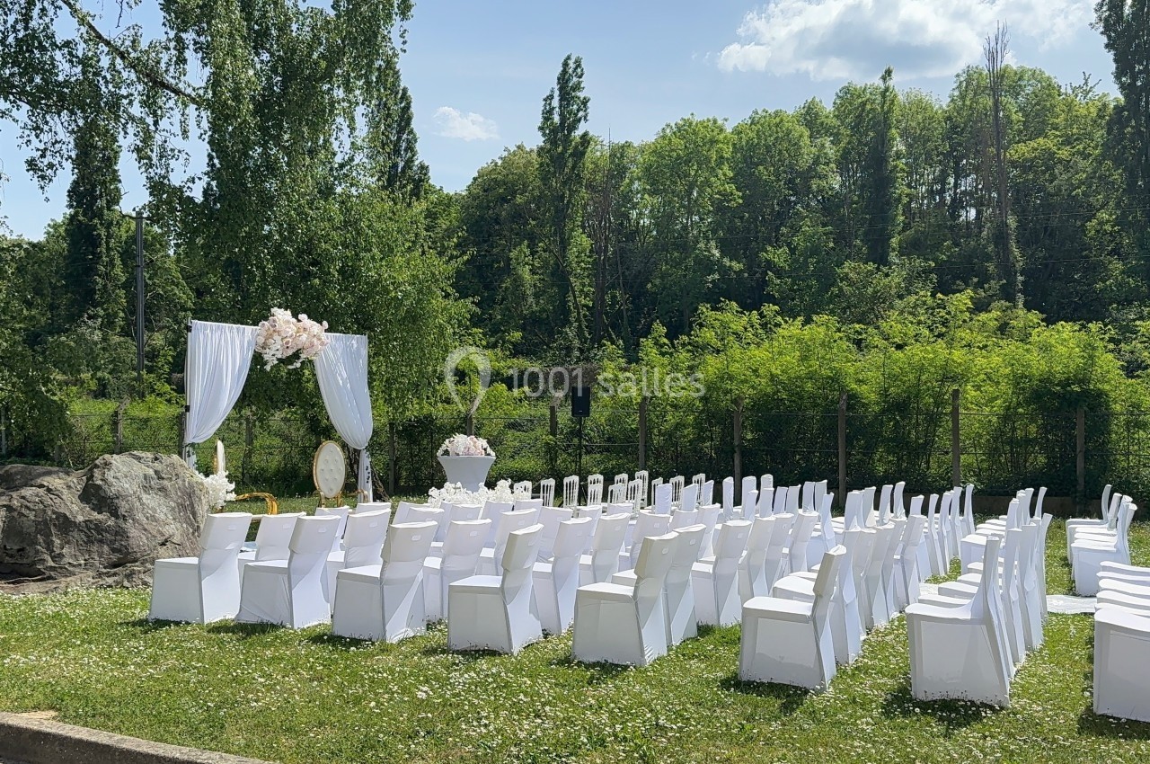 Chaises blanches alignées en extérieur devant une arche décorée de fleurs, entourées de verdure et d'arbres.