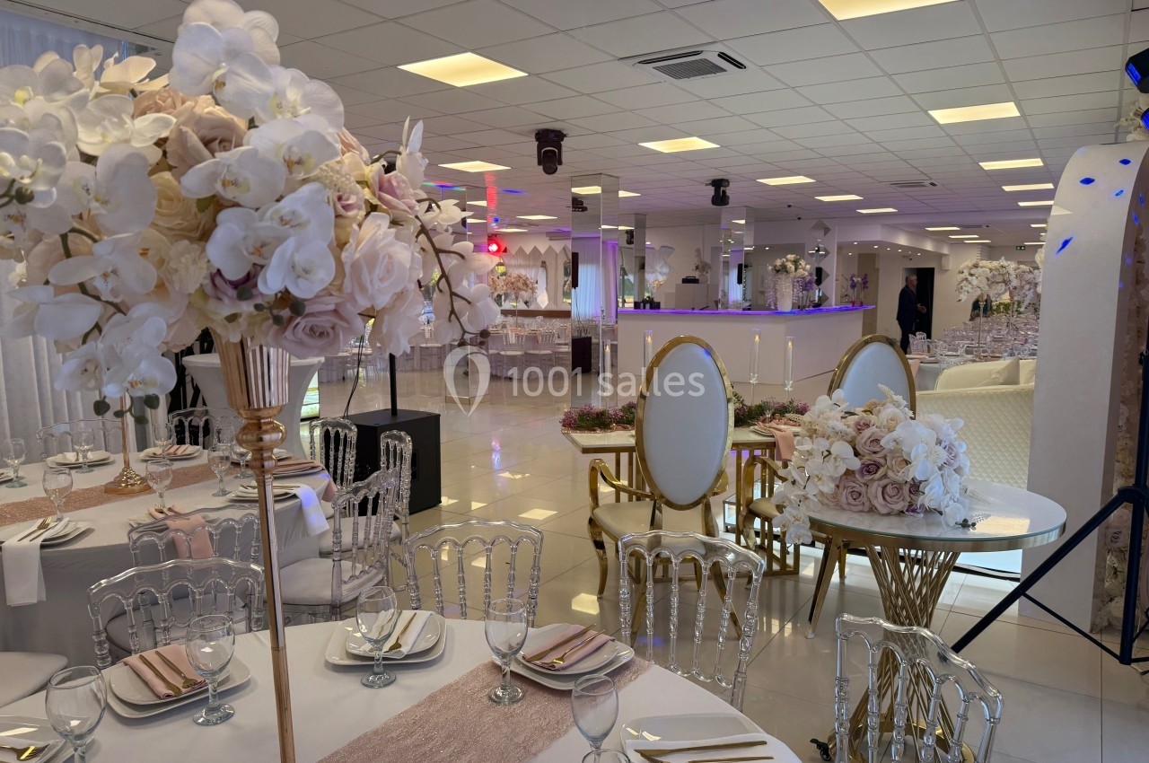Salle de réception élégante avec tables décorées de fleurs blanches et roses, chaises transparentes et éclairage chaleureux.