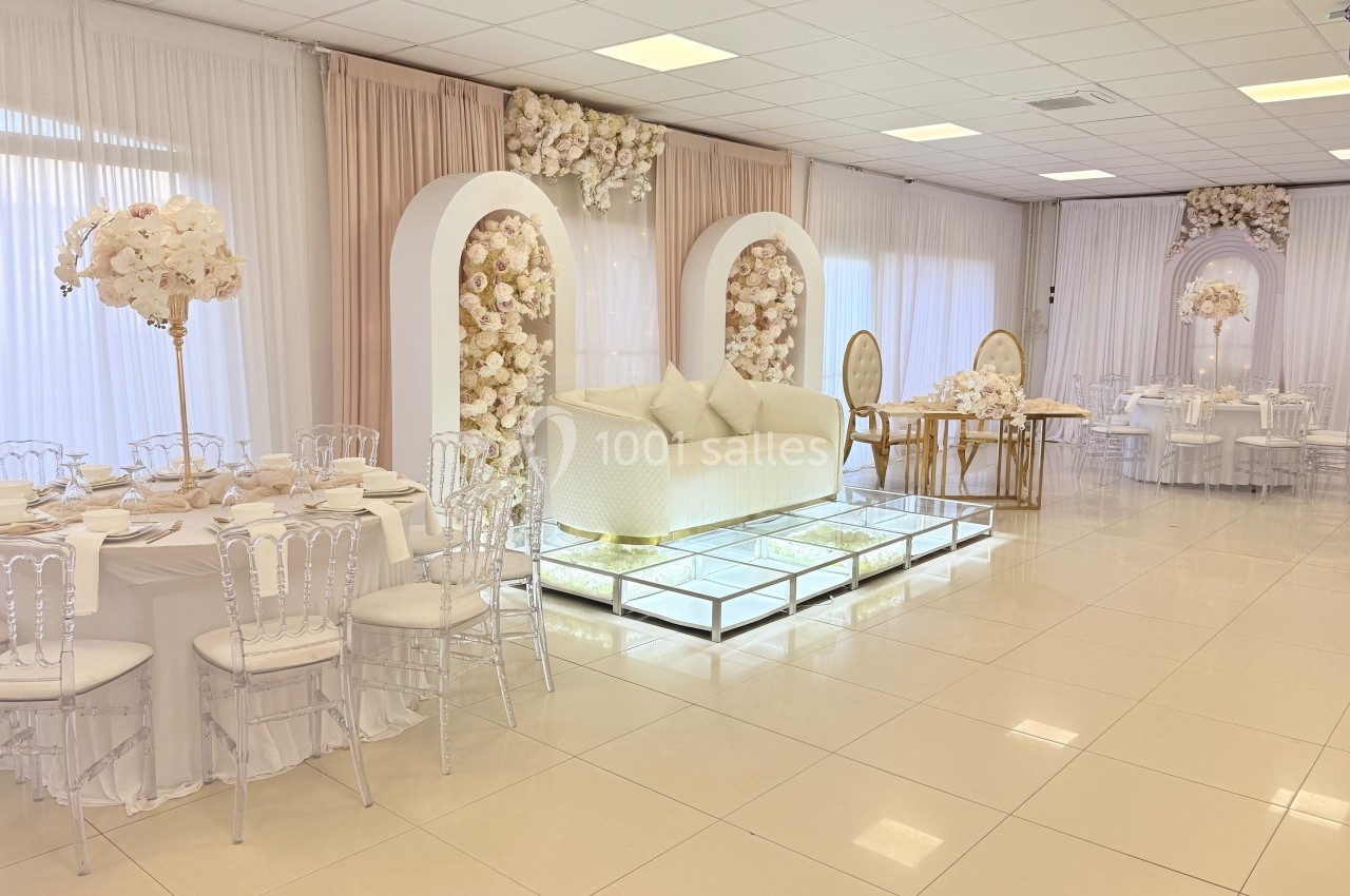 Salle de réception lumineuse avec décor floral blanc et beige, tables dressées et canapé sur estrade vitrée.