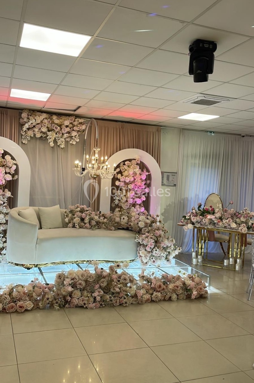 Décoration de salle avec canapé blanc, fleurs roses et blanches, arches fleuries et table ornée dans un cadre élégant.